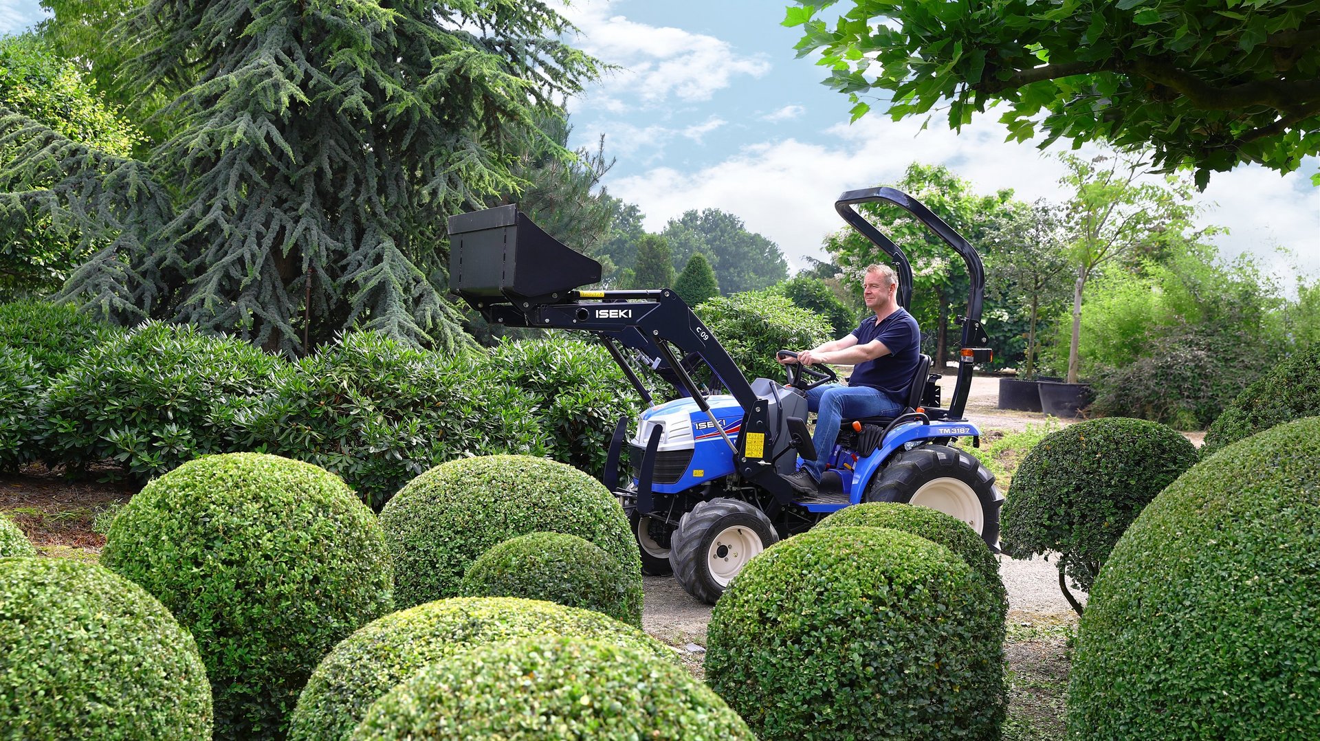 Mann transportiert Pflanzen mit Hilfe des Frontlader eines ISEKI-Traktors durch einen Garten..